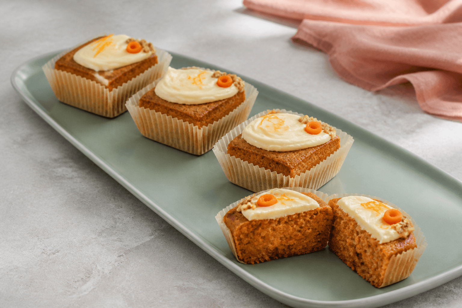 Tre hele firkantede gulerodsmuffins og én åbnet muffin på havgrønt fad med lav citrus-ostecreme, gulerodskrølle og nøddeknas