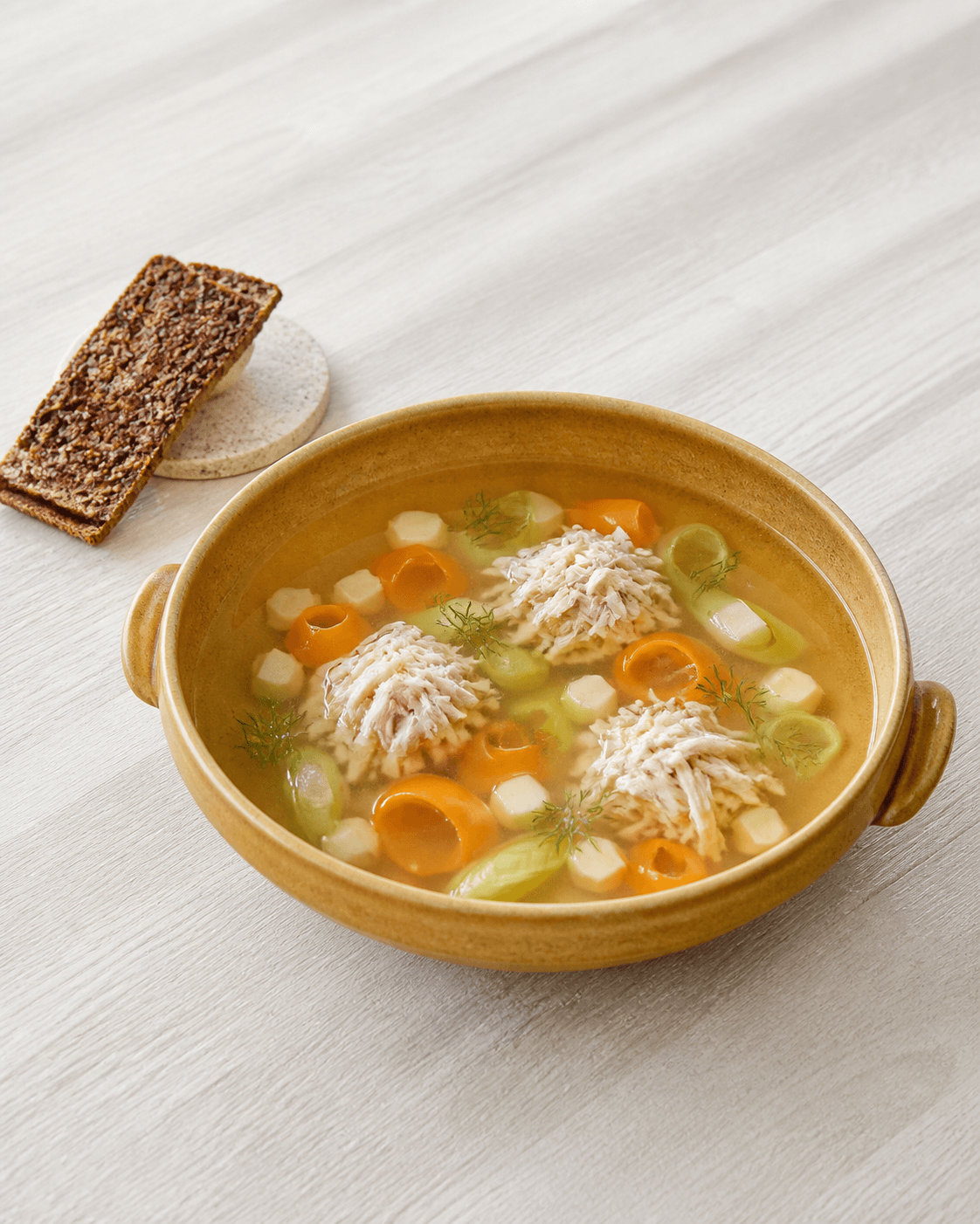 Klar gylden kyllingesuppe i ravfarvet skål med tre bunker trevlet kylling, gulerodsribbons, porre, selleri, dild og kerneknaekbroed