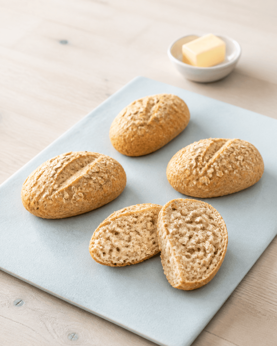 Tre hele aflange glutenfri skyrboller og én delt på lyseblå bageplade med havre, solsikkekerner og en lille smørskål