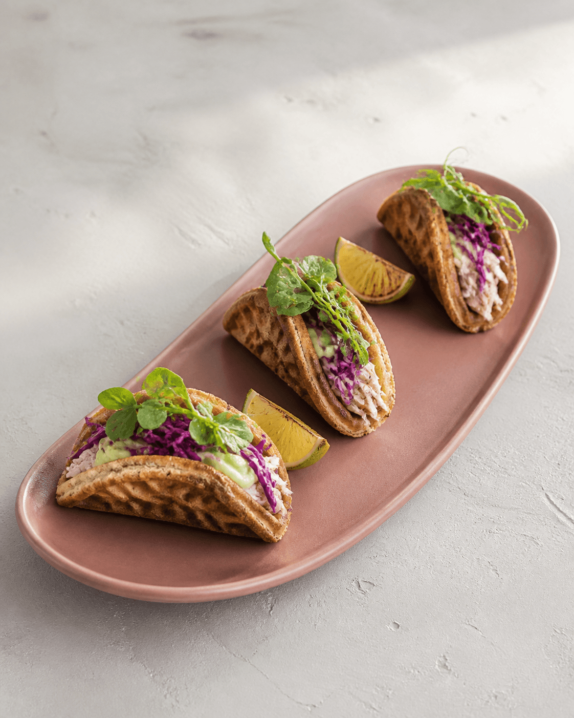 Tre foldede runde rugvaffeltacos på støvet rosa fad med trevlet kylling, avocadomousse, cider-rødkål, ærteskud og ristede limebåde