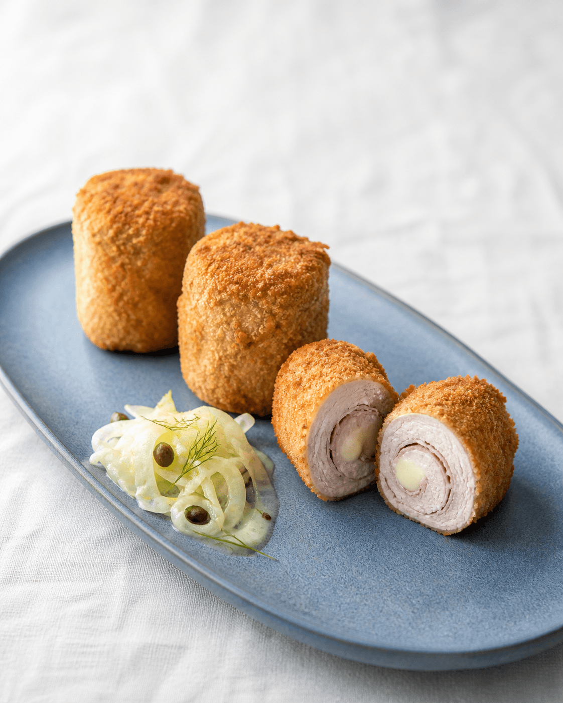 To hele og én skåret paneret kalve-cordon-bleu-roulade på støvetblåt fad med synlig skinke, gruyère, fennikel og kapers