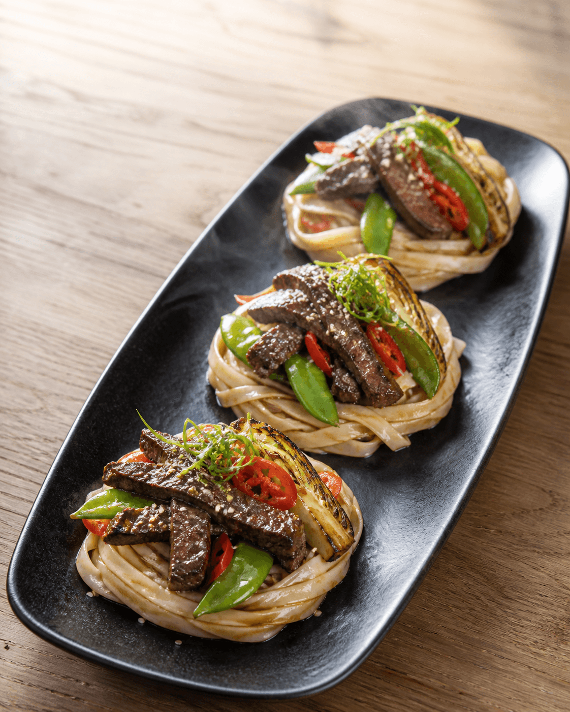 Tre separate reder af brede risnudler på sort auberginefarvet fad med gennemstegt oksekød, grillet spidskål, sukkerærter og rød peber