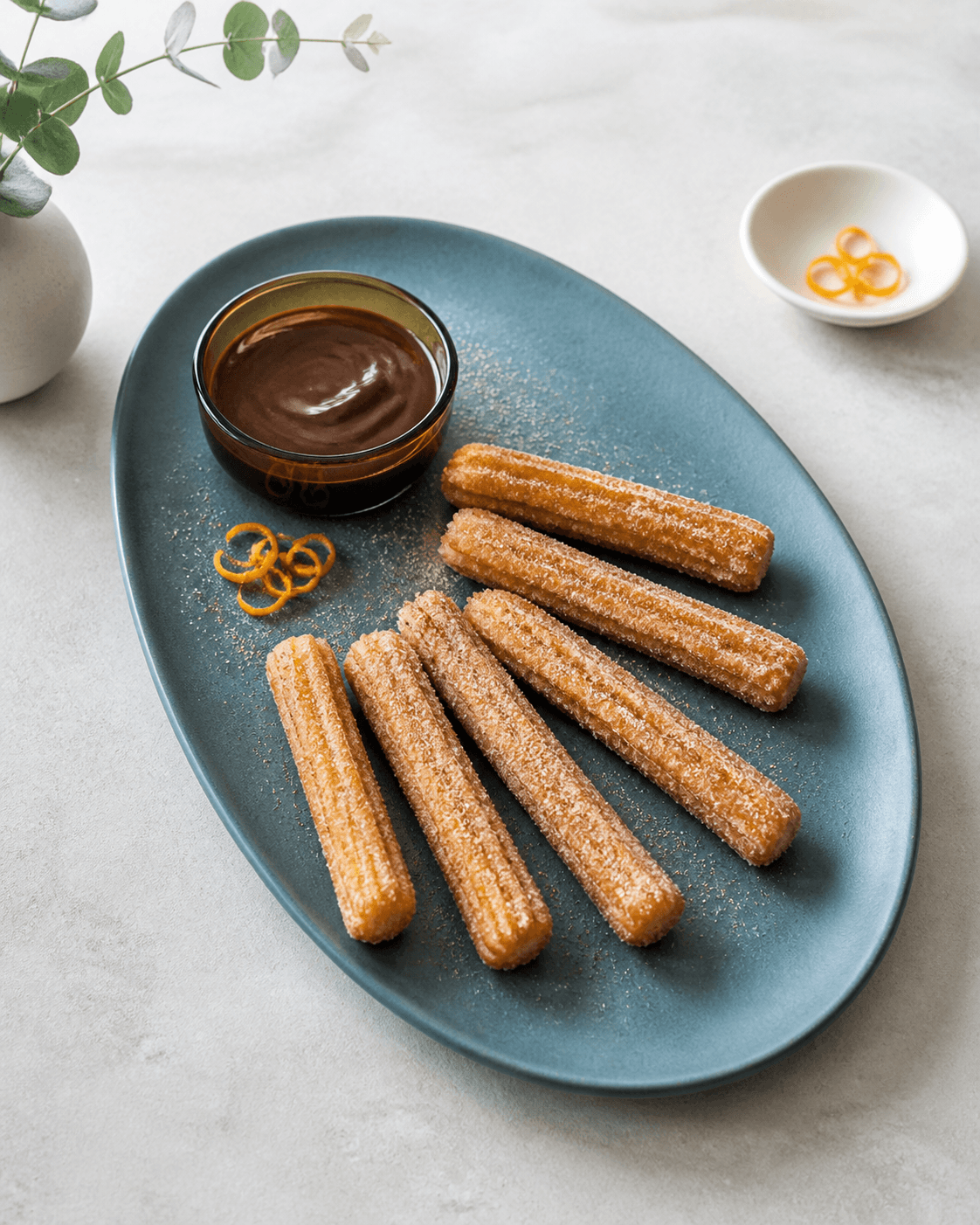 Syv lige sprøde churros i vifte på petrolblåt fad med mørk chokolade, appelsinskal og kanelsukker