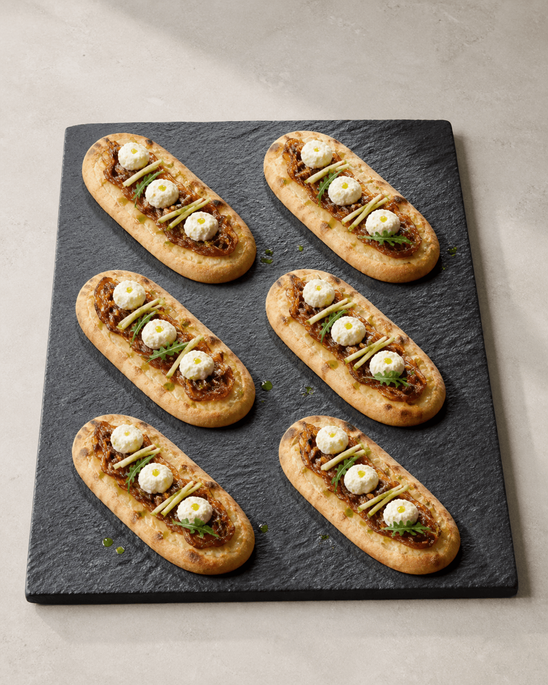 Seks aflange ovale pizzette på sort stenfad med karamelliseret løg, tre fetaprikker, grønne æblestave og rucola