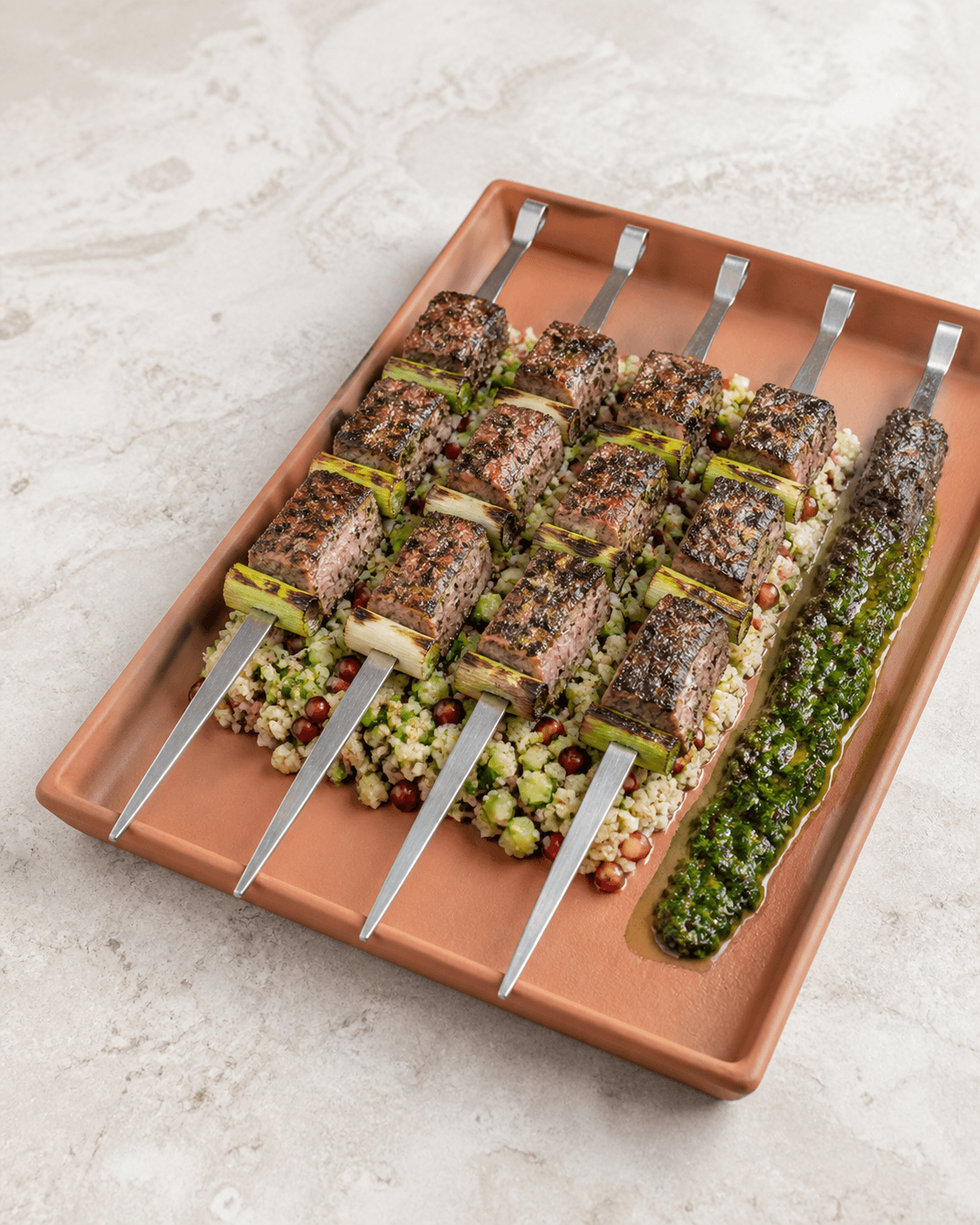 Fem flade metalspyd med tre grillede lammestykker og forårsløg på terracottafad over citronbulgur med agurk, granatæble og chimichurri
