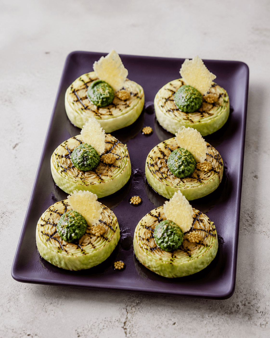Seks runde grillede spidskålsrosetter på auberginefarvet fad med persille-valnøddepesto, parmesancrisp og syltede sennepsfrø