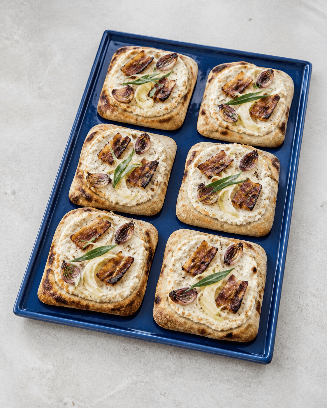 Seks rektangulære pizza bianco-pizzette på koboltblåt fad med ricotta, sprød bacon, rødløg, syltet fennikel og estragon