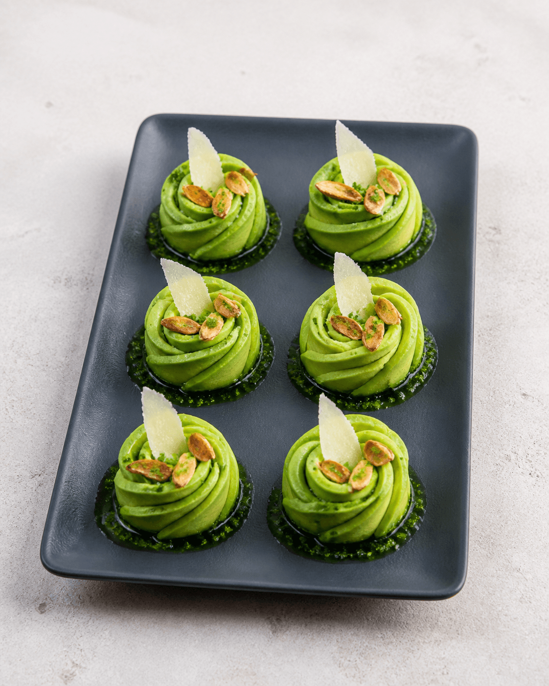 Seks avocadoroser på koksgråt fad med parmesansejl, tre ristede mandler, purløg-persilleolie og citronsalt
