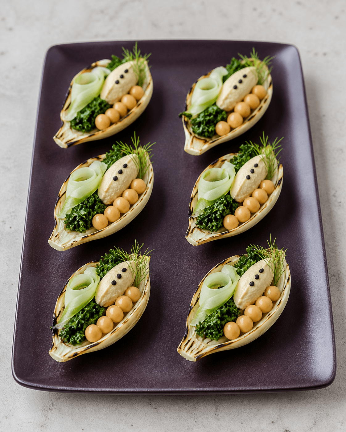 Seks grillede fennikelbåde på auberginefarvet fad med fem kikærter, selleribånd, grønkål, citronhummus, tre sorte sesamfrø og fennikeltop