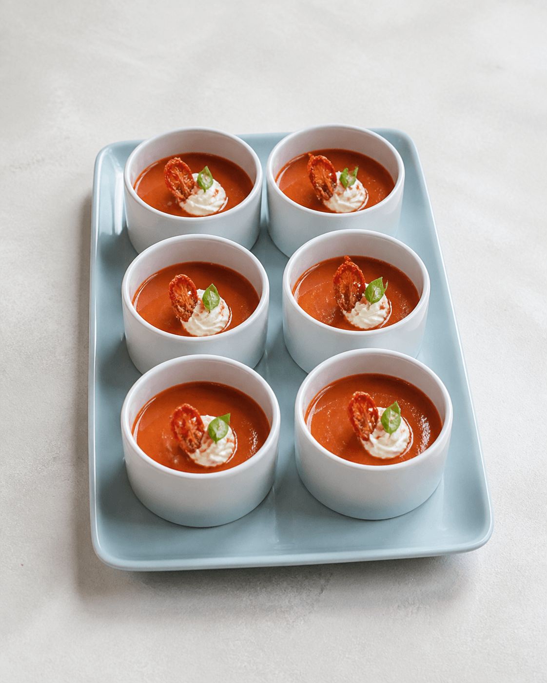 Seks små hvide kopper med bagt tomatsuppe på isblåt fad, ristet hvidløgsskyr, sprød tomatskindsspiral, basilikum og peberfrugtolie