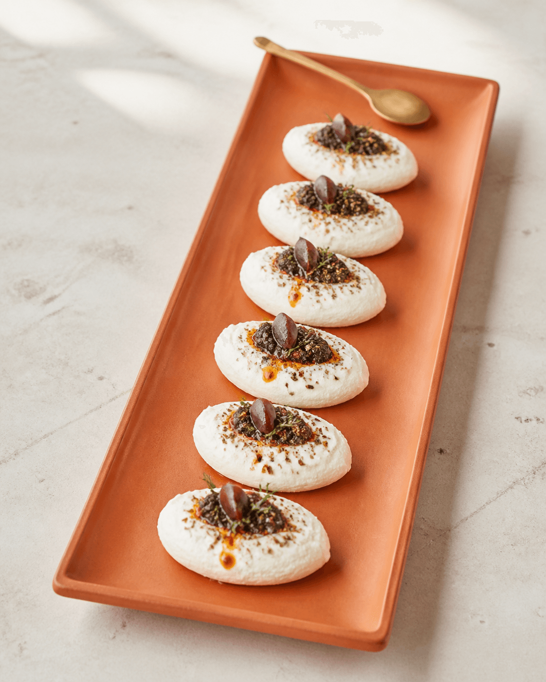 Seks ovale labneh-queneller på brændt orange fad med oliventapenade, za'atar, timian og chiliolie