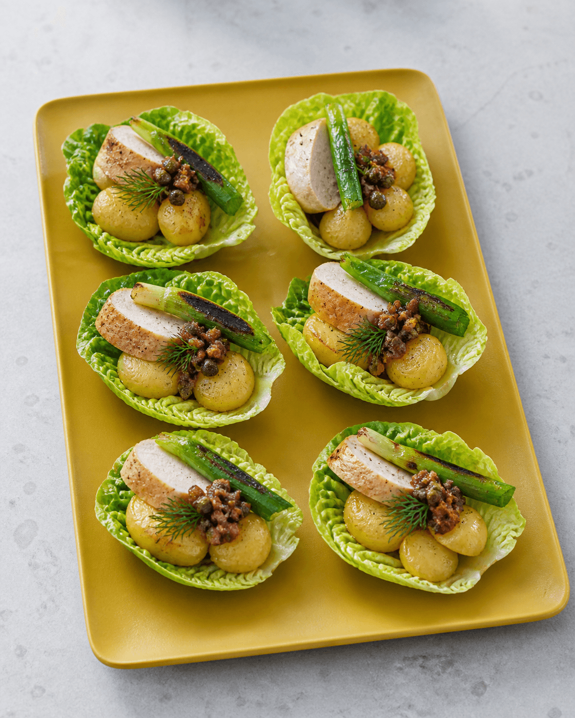Seks hjertesalatsskåle på sennepsgult fad med stegte kartofler, kylling, grillet forårsløg og bacon-kapers-dressing