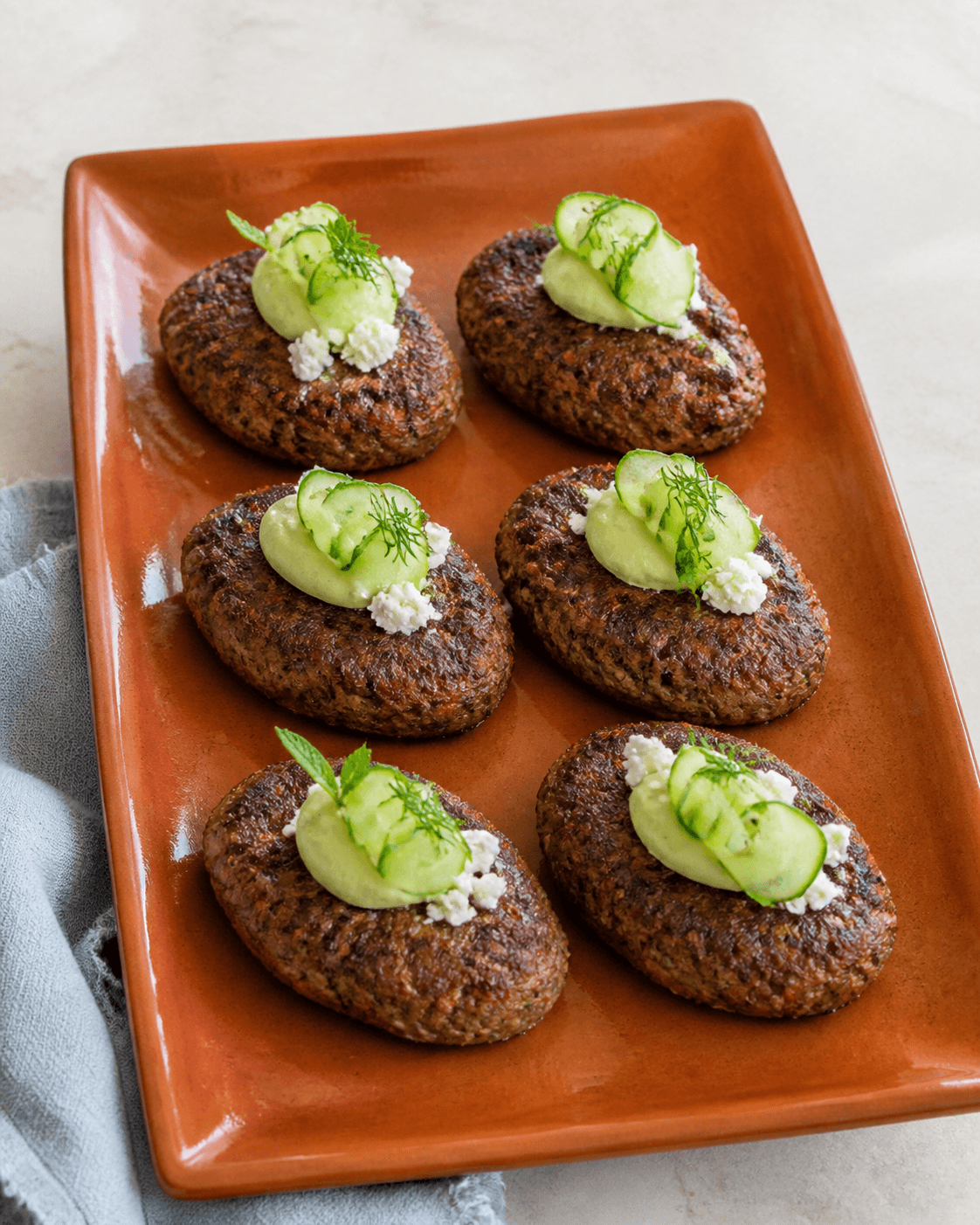 Seks ovale lammefrikadeller på terracottafad med avocadocreme, agurk, feta og dild