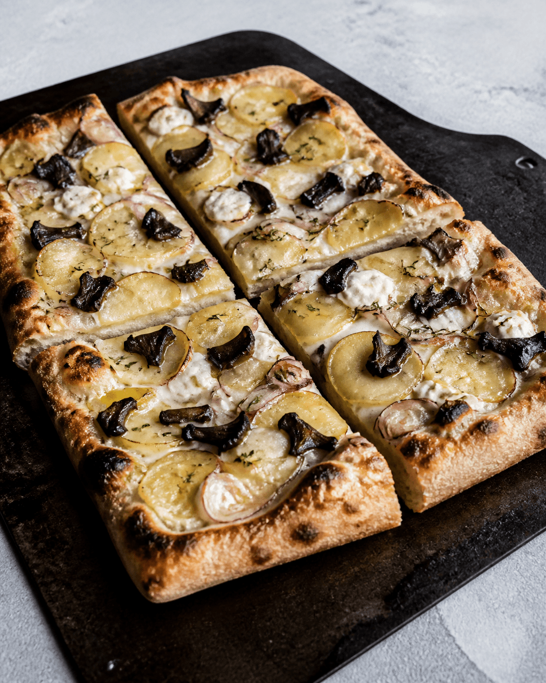 Rektangulær hvid pizza i fire stykker med kartoffelskiver, rødløg, trompetsvampe, mascarpone, ricotta og timian