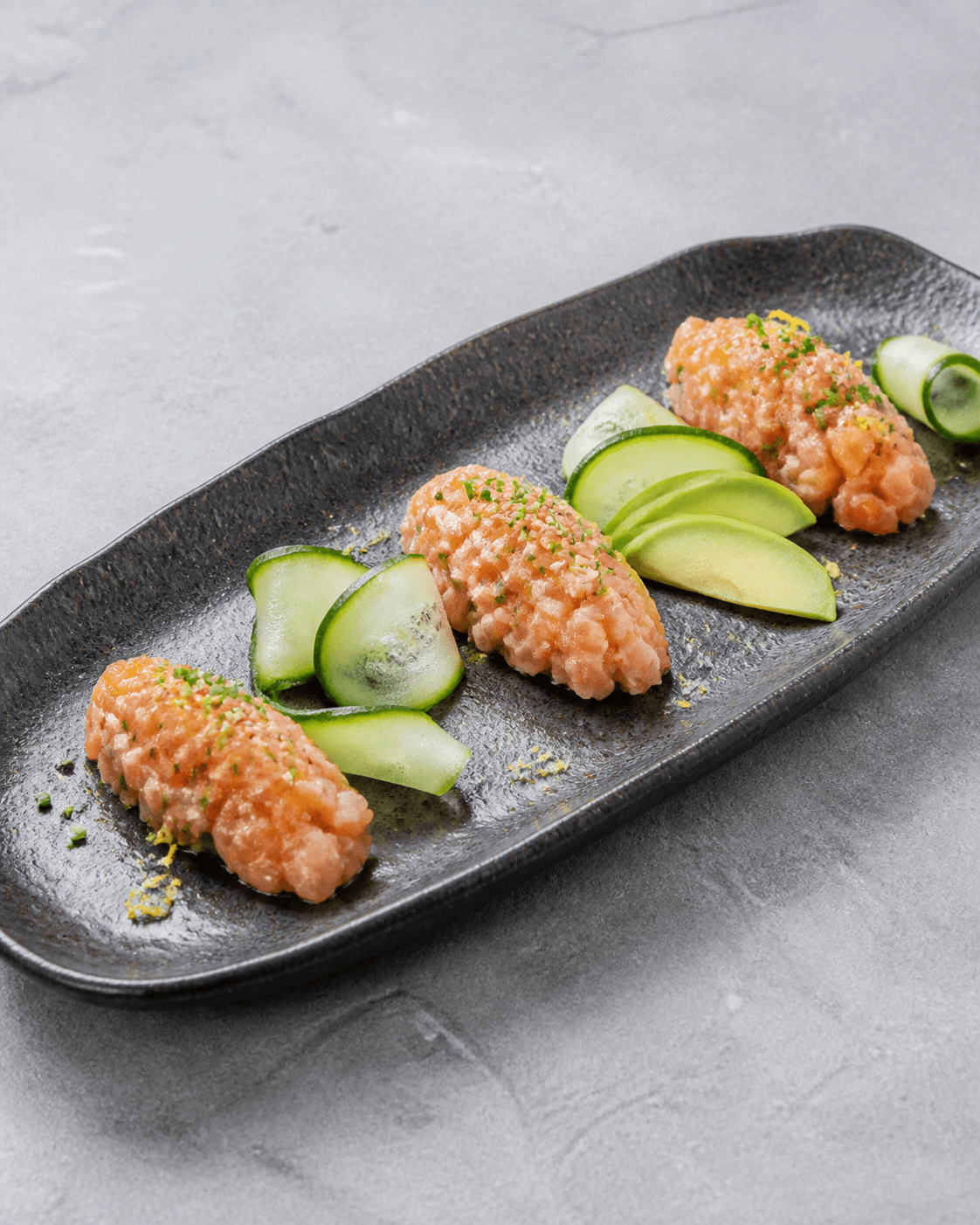 Tre kolde queneller af fintskåret rå laks med avocadofaner, agurkebånd, purløg, citron og sesam på mørkt aflangt fad