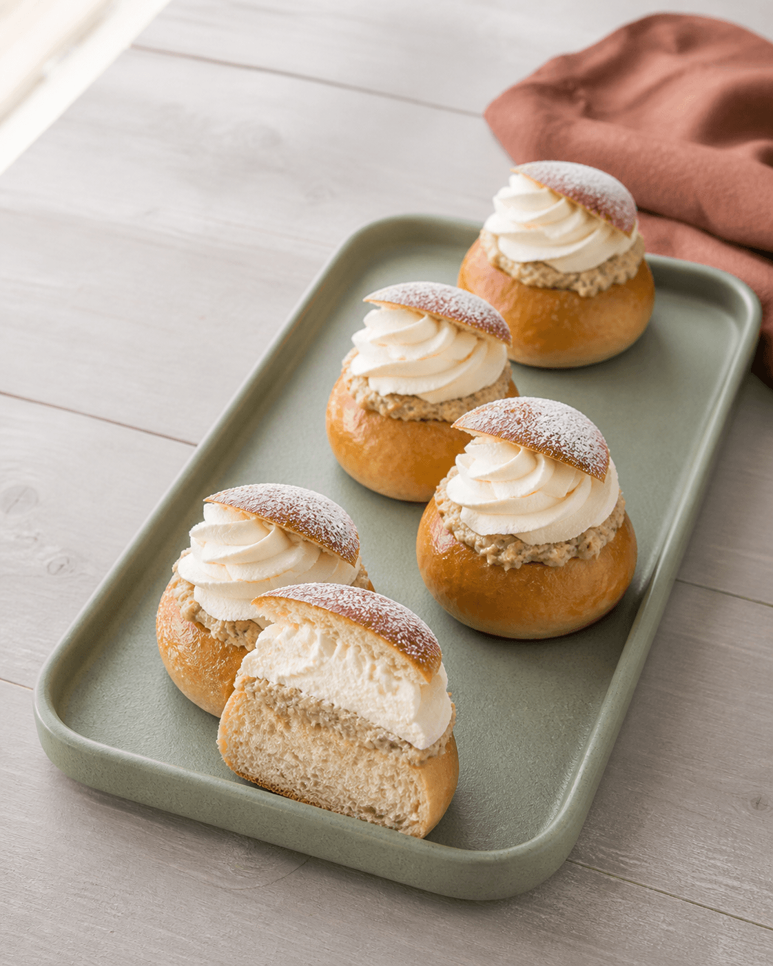 Fem svenske semlor med gyldne kardemommeboller, groft mandelfyld, flødeskum og flormelis på salviegrønt bagefad