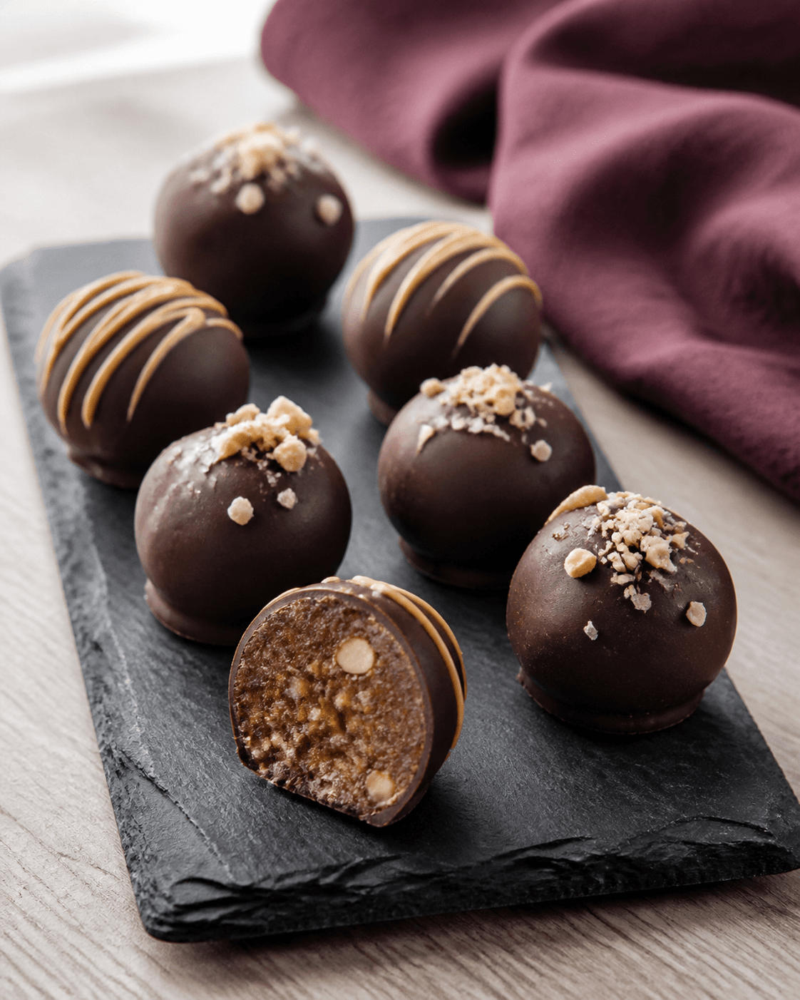 Syv mørke chokoladekugler med dadel-peanutbutterfyld, peanutstriber, hakkede peanuts og flagesalt på sort skiferfad