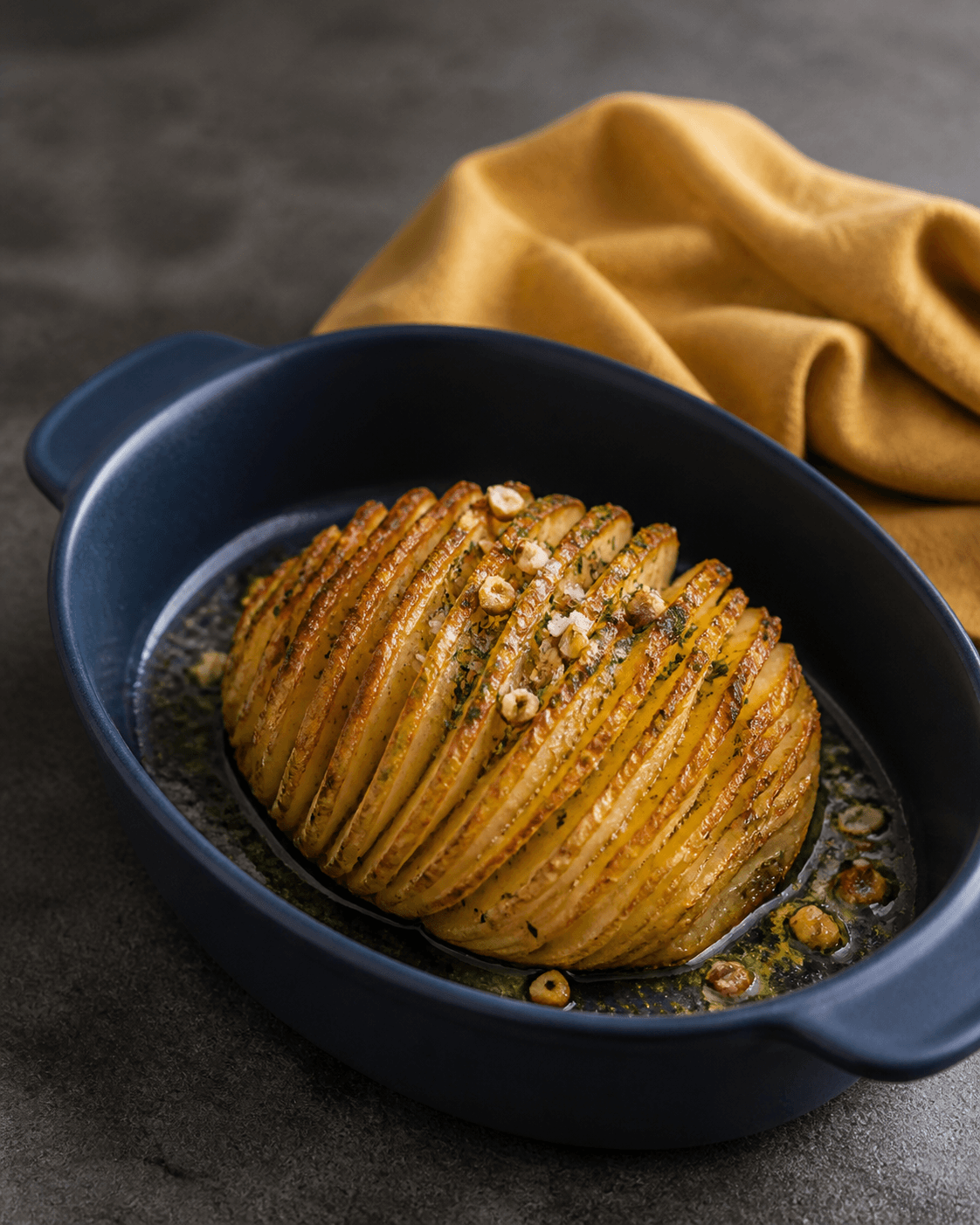 Hel rund hasselback-knoldselleri med dybt gyldne skiver, persille-tarragonsmør og ristede hasselnødder i mørkeblåt ovalt fad