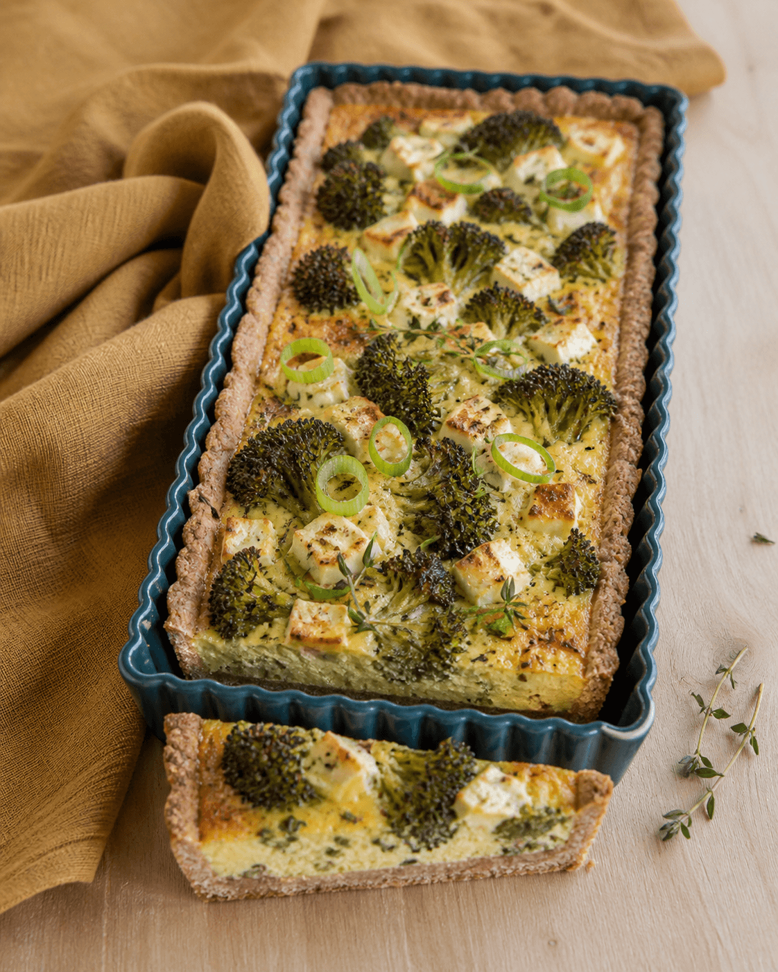 Rektangulær fuldkornstærte med helt sat gyldent æggefyld, broccoli, pasteuriseret feta, forårsløg og timian i mørk petroleumsfarvet form