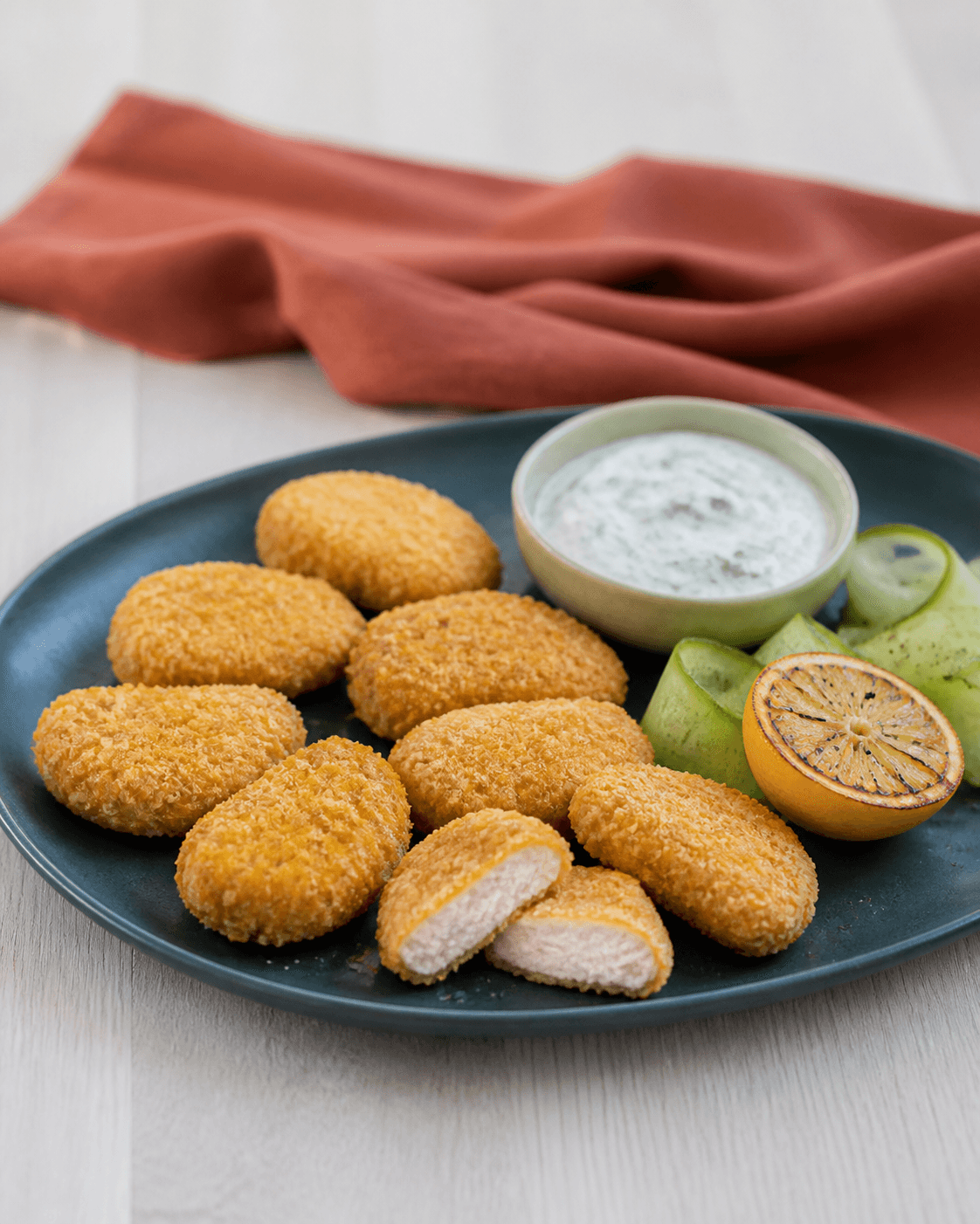 Otte ens ovnbagte kyllingenuggets med tynd gylden panko-cornflakeskorpe, én gennemskåret med helt hvid kerne, yoghurtdilddip, agurk og grillet citron på mørkt teal fad