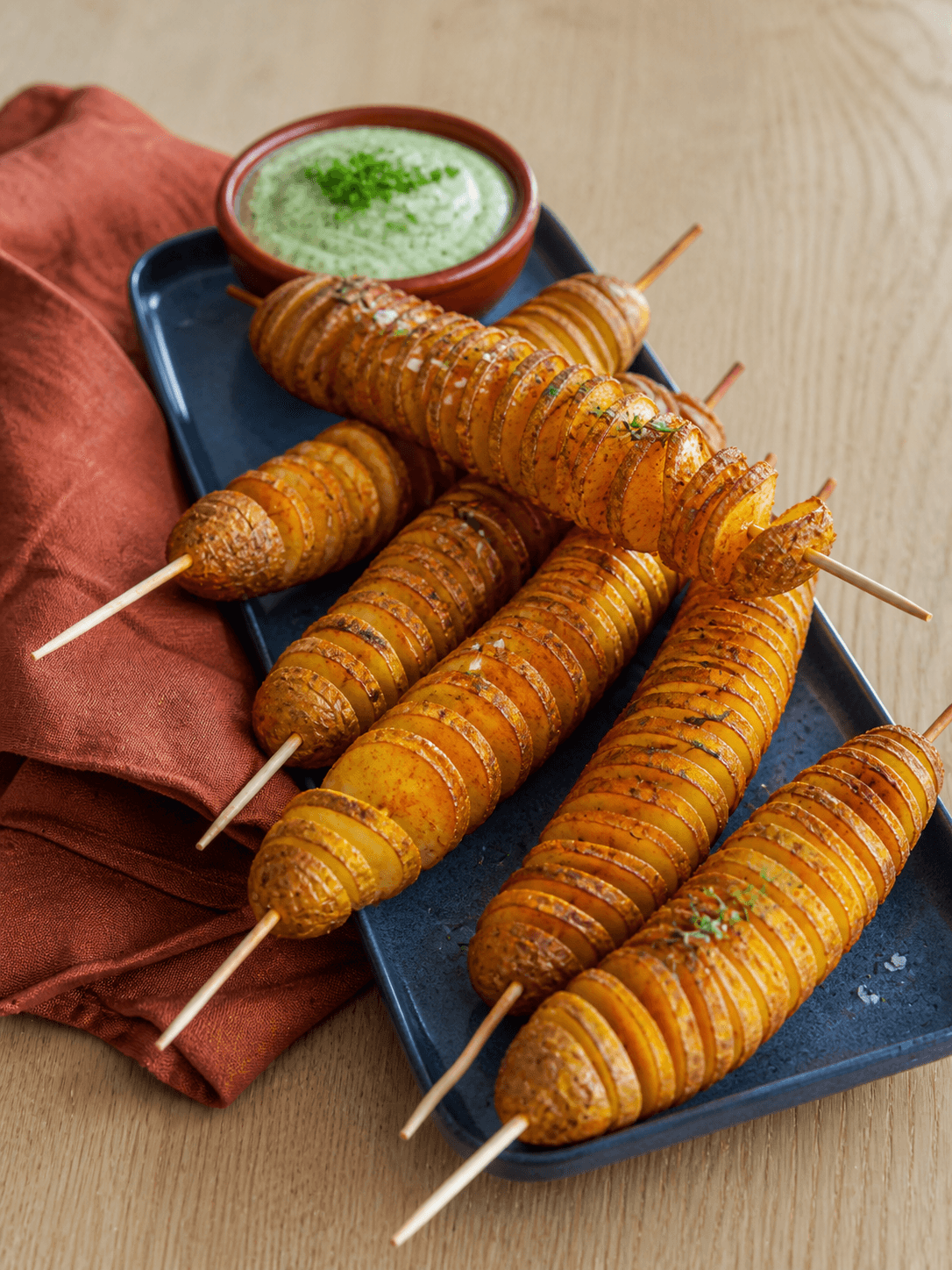 Fem hele sprøde kartoffelspiraler på træspyd med paprika, rosmarin og flagesalt på smalt mat indigoblåt fad med grøn vegansk purløgsdip i terrakottaskål