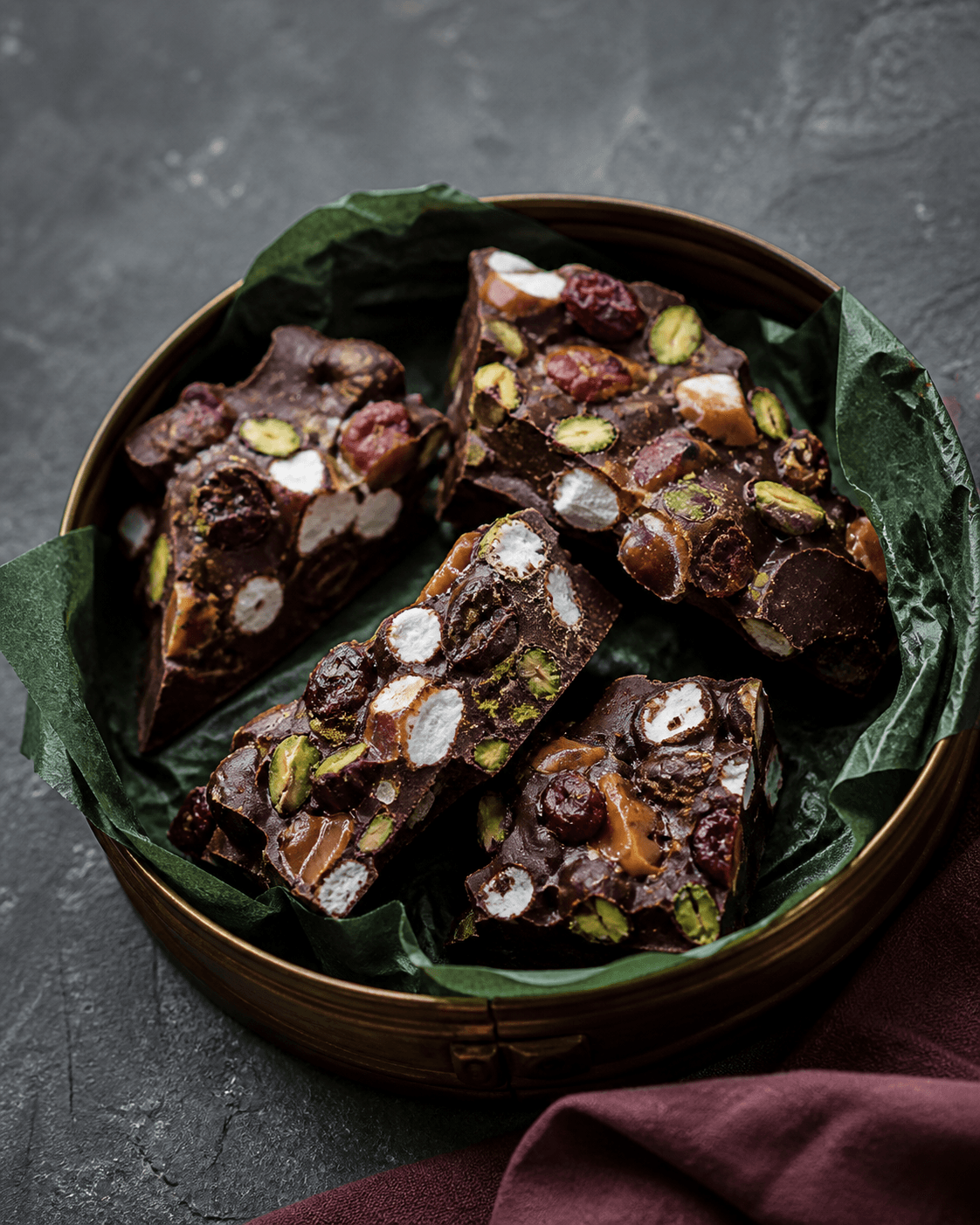 Uregelmæssige mørke rocky-road-stykker med skumfiduser, saltkaramel, pistacier og tørrede tranebær i åben rund messingdåse med mørkegrønt papir