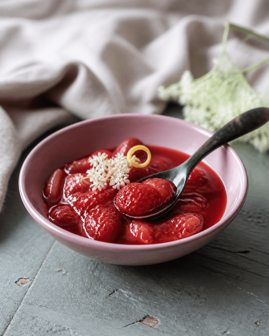 Dyb rubinrød jordbær-rabarberkompot med bløde frugtstykker i mat støvet rosa skål, mørk ske med jordbær, hyldeblomst og citronskal