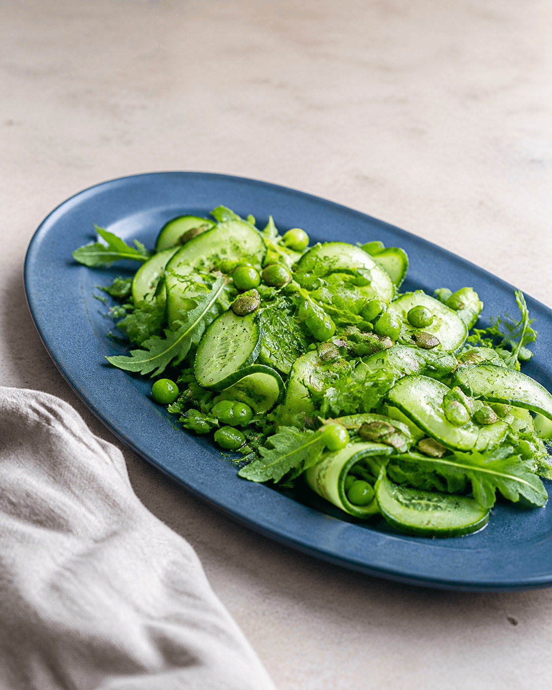 Kold sprød agurkesalat med halve skiver og løse bånd, rucola, grønne ærter, dild, græskarkerner og citron-honningvinaigrette på langt mat koboltblåt fad