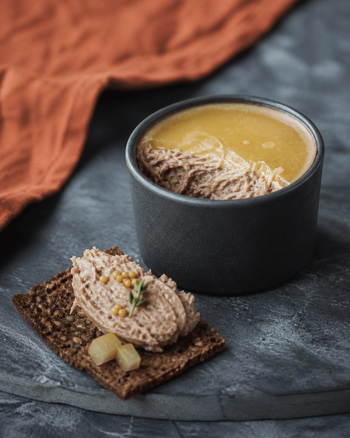 Kold grov anderillette i lille mat koksgrå krukke med tynd ravfarvet fedtforsegling og åbent felt med tydelige fibre samt en quenelle på mørkt rugknækbrød