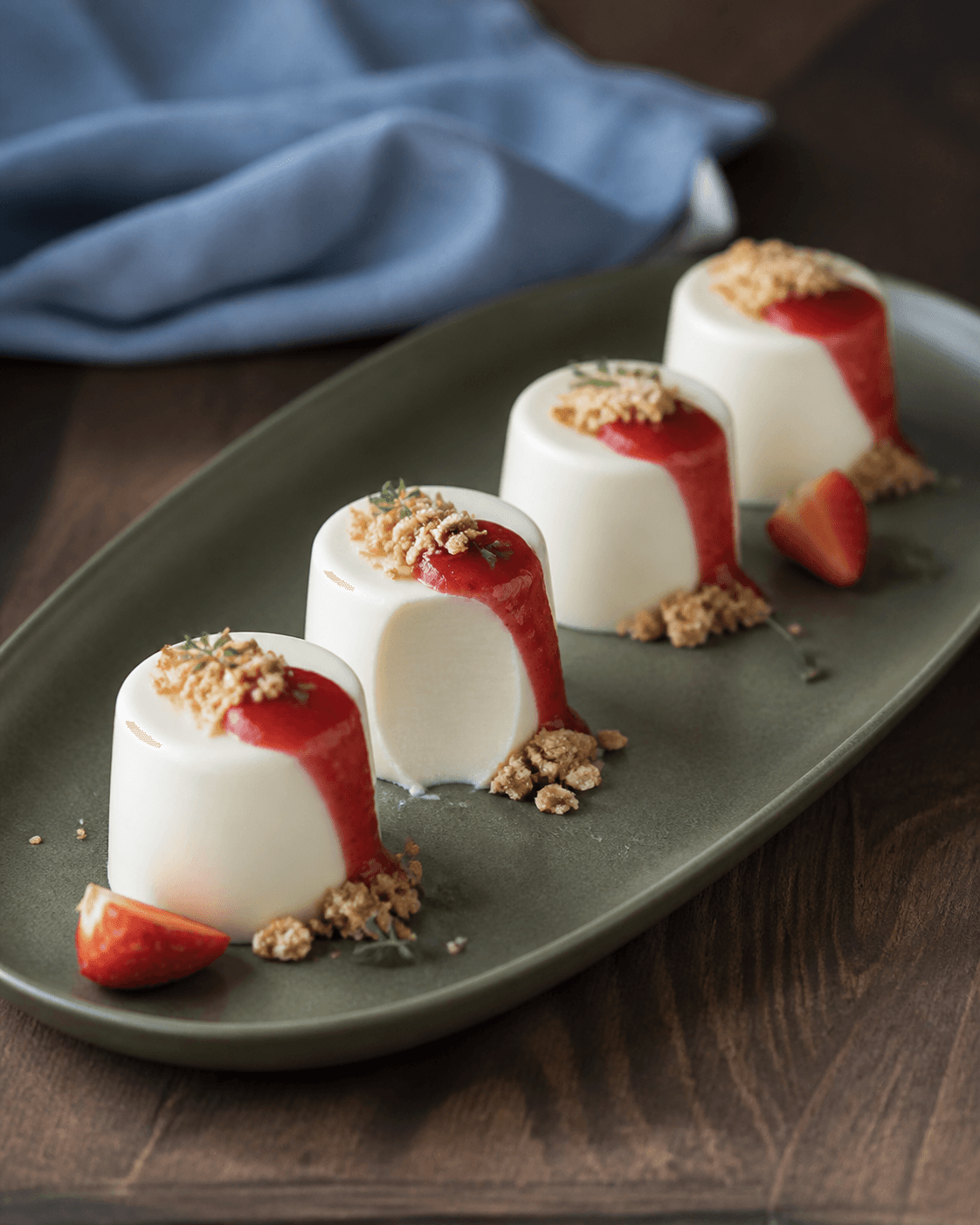 Fire fritstående hvide chokolade-panna cotta med jordbær-rabarberbånd og gylden crumble på langt salviegrønt fad, én med rent ske-snit