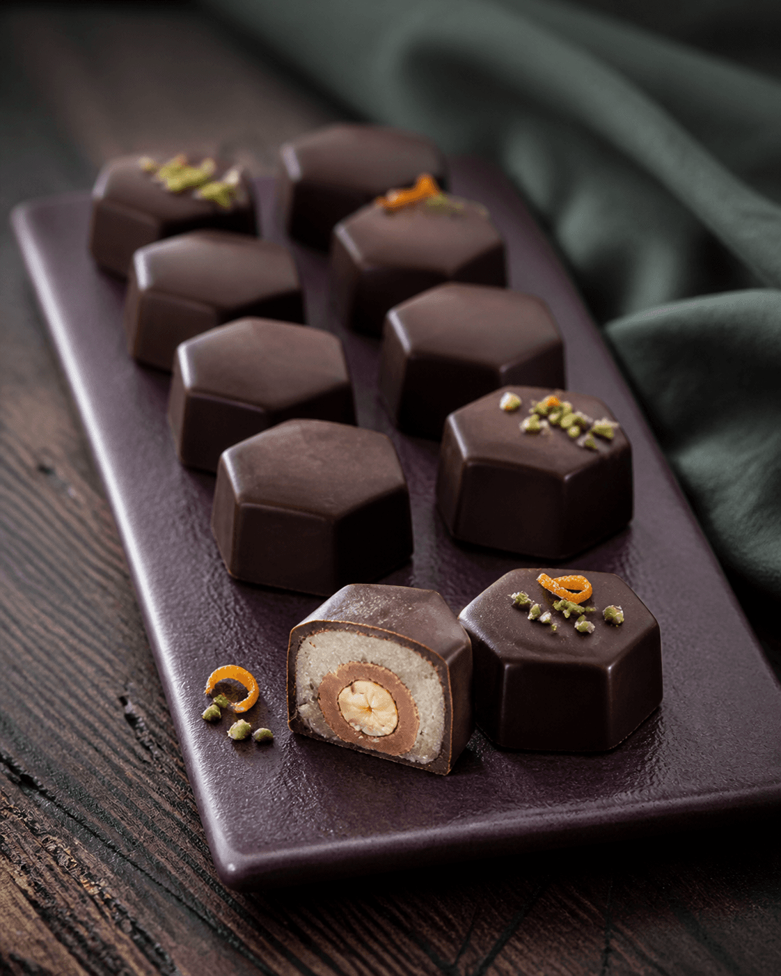 Facetterede mørke Mozart-konfektstykker i to rækker på auberginefarvet fad, én skåret med hasselnød, nougat, pistaciemarcipan og tynd chokoladeskal