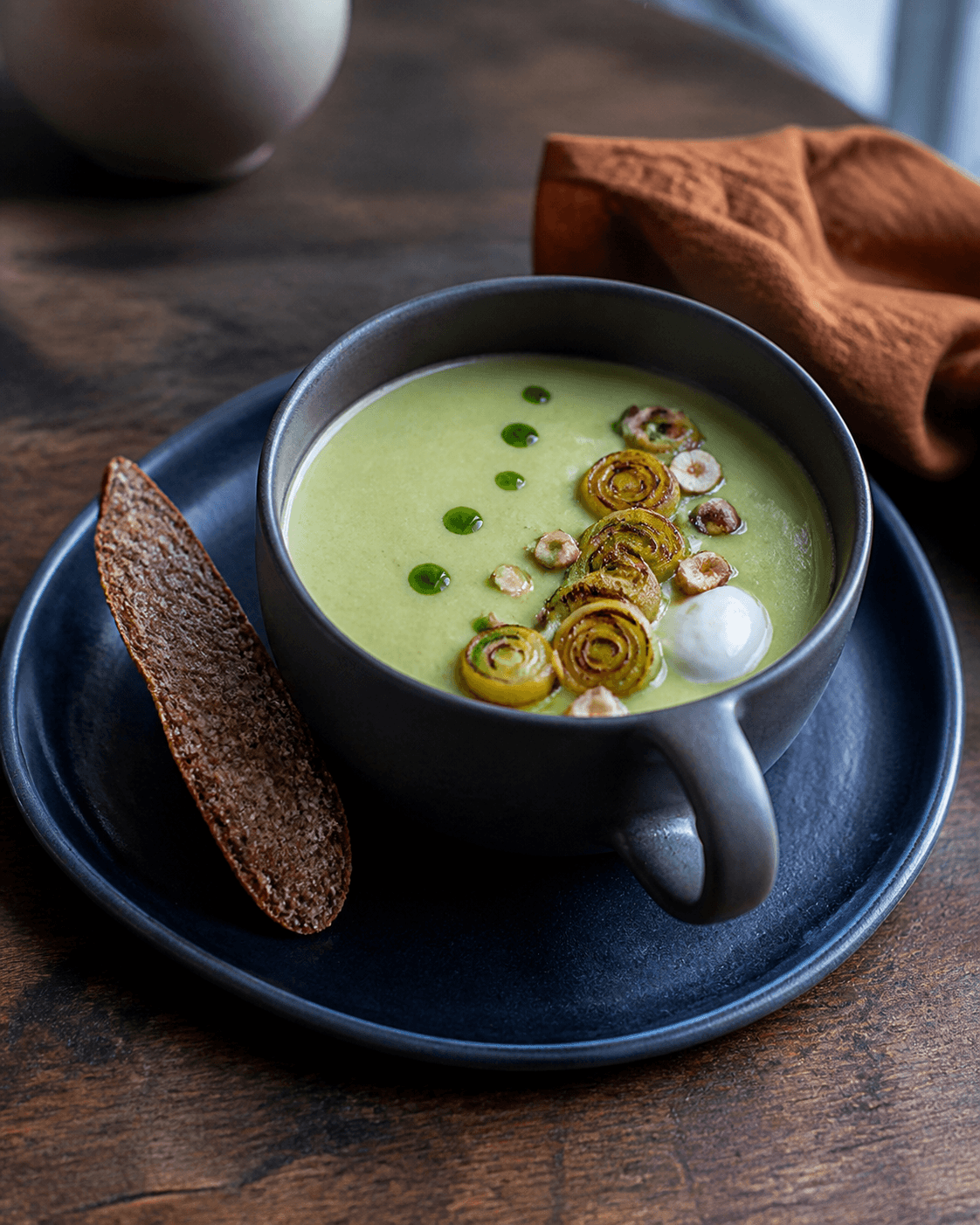 Palegrøn porre-kartoffelsuppe i mat koksgrå kop på midnatsblå tallerken med brunede porreringe, hasselnød, purløgsolie, syrnet fløde og rugknæk