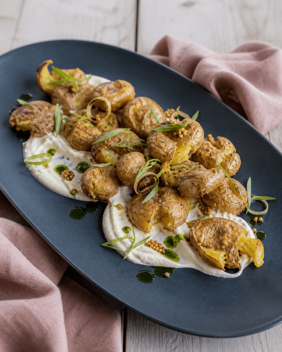 Sprøde knuste nye kartofler i en bue på langt midnatsblåt fad med pisket feta-citronsauce, estragon, sennepsfrø, grøn olie og grillede løgringe
