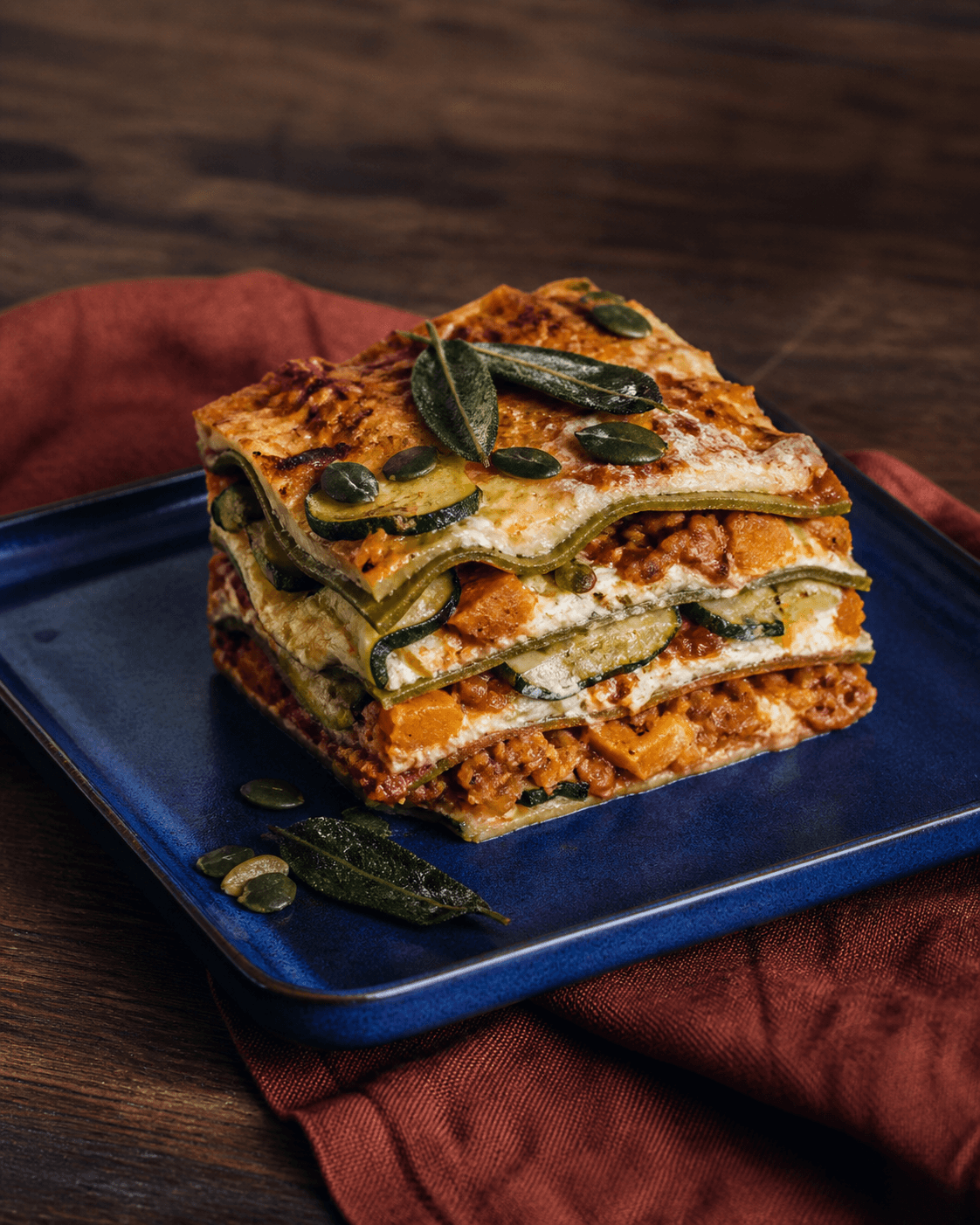 Høj squashlasagne med grøn pasta, tomat-linseragù, orange græskar, zucchini og sprød salvie på koboltblå plade
