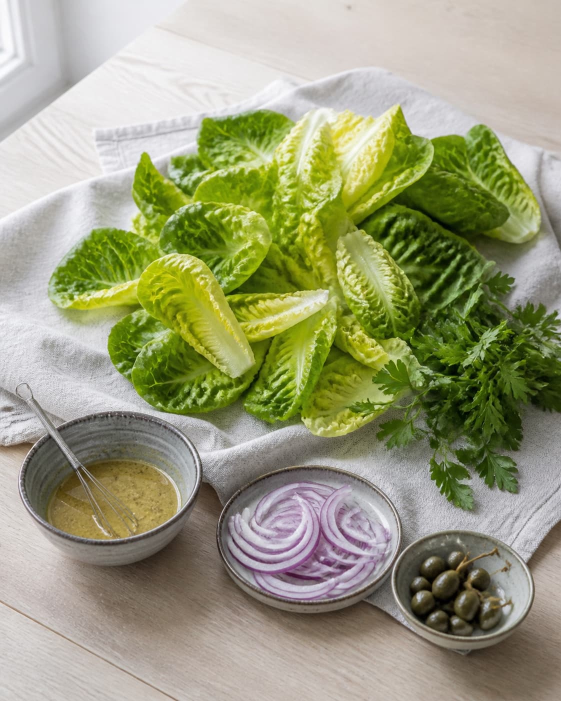 Tør romainesalat og persille ligger på lyst klæde med citron-dijonvinaigrette, rødløg og kapers i små skåle