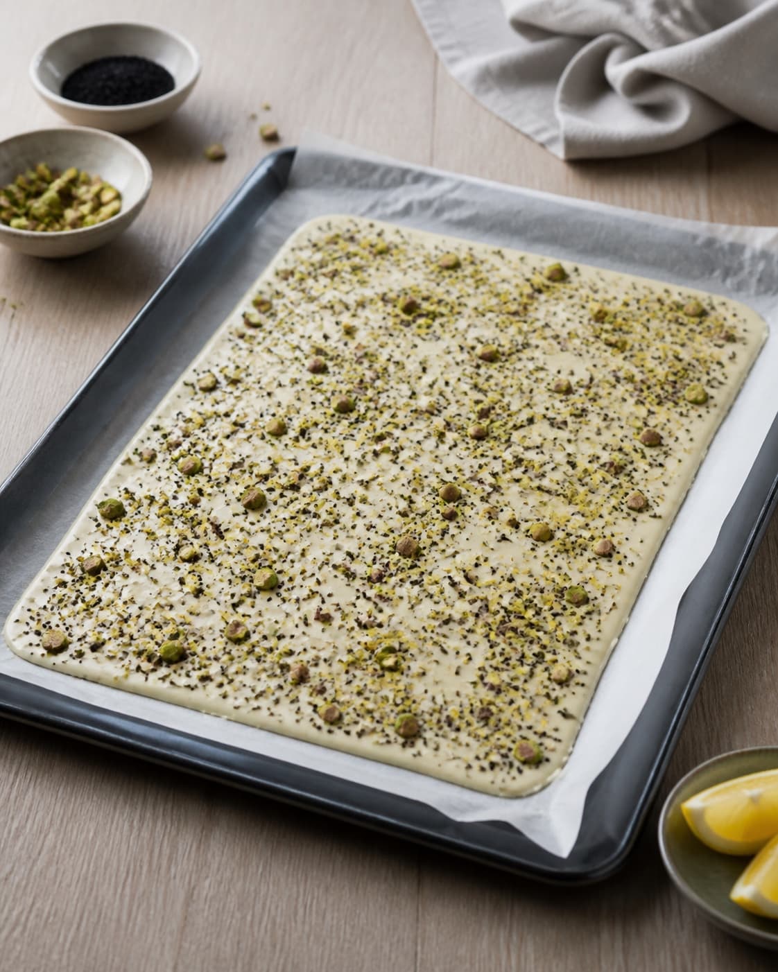et ens tyndt lag blank hvid chokolade på bagepapir drysset jævnt med grøn pistacie, citron og sort lakrids; kontroltegnet er laget er ens tykt, og toppingen fordeles uden bare eller overfyldte felter
