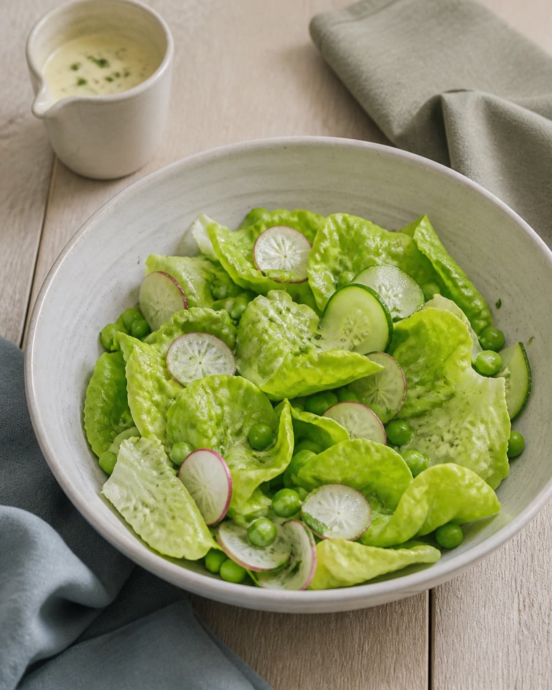 sprøde smørsalatblade, agurk, radise og ærter ligger i en skål med dressing i en separat lille kande klar til sparsom vending; kontroltegnet er salaten er blankt men let dækket, bladene stadig sprøde og der ligger ingen dressingpøl i bunden