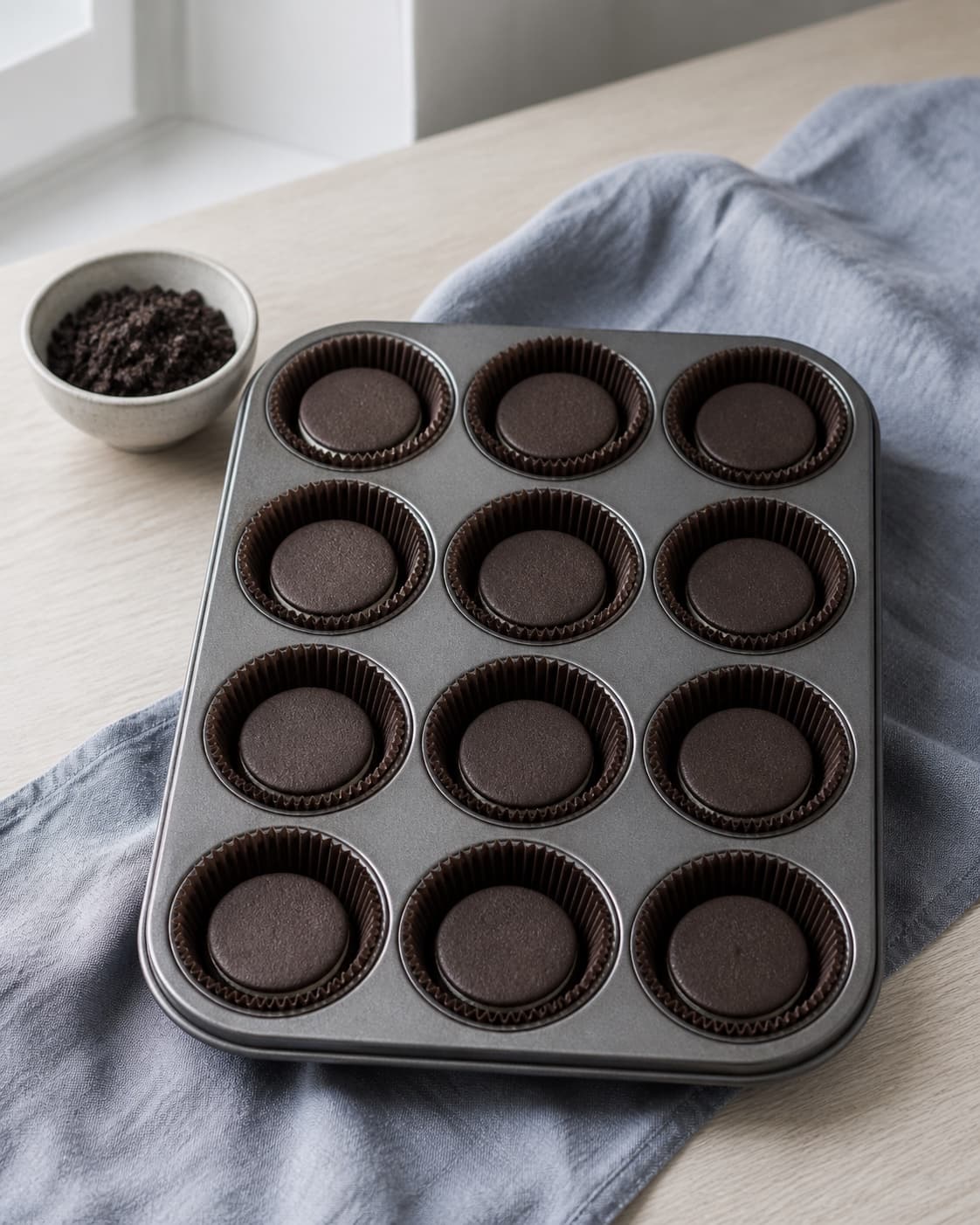 tolv ens mørke papirsforme står i fast muffinform med én ubrandet chokoladesandwichkiks plant i hver bund; kontroltegnet er hver form har præcis én hel kiks i bunden, og finishsmulderet er groft uden pulver
