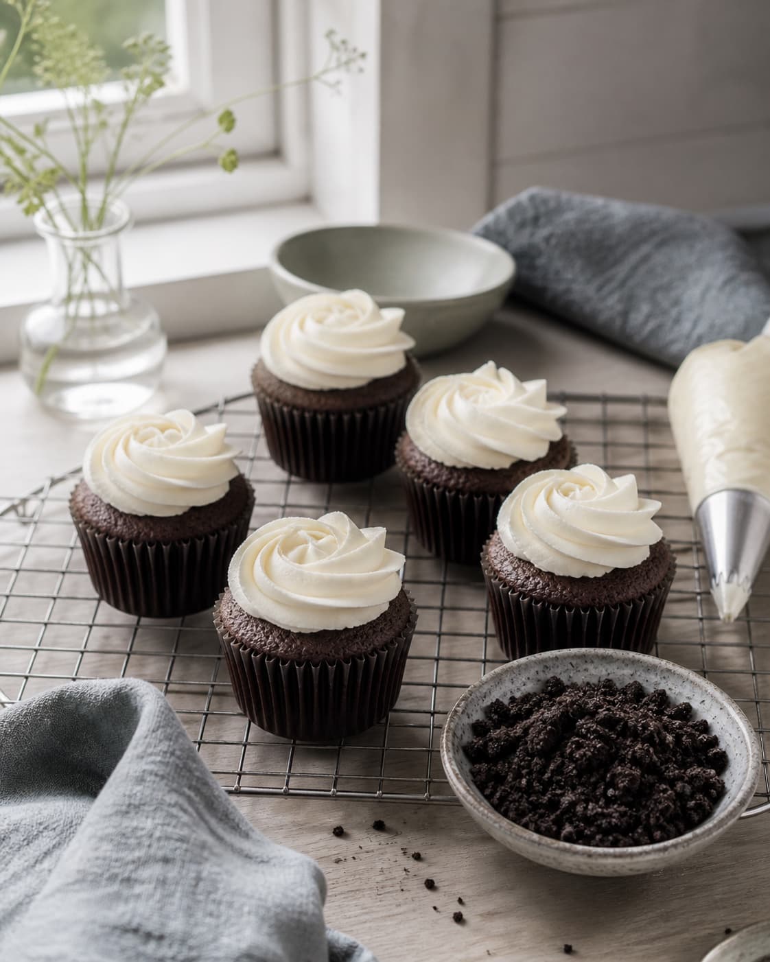 kolde kakaocupcakes får moderate hvide vaniljerosetter fra sprøjtepose og grove ubrandede mørke kiksesmulder drysses separat; kontroltegnet er hver cupcake har en ren stabil vaniljeroset og grove sprøde mørke smulder uden smeltet creme