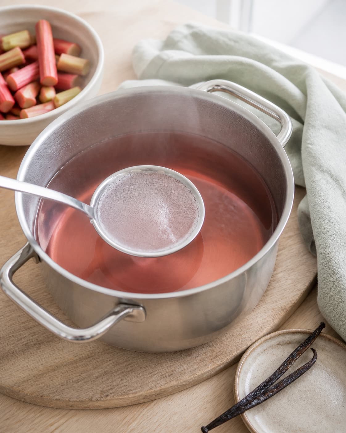 blank klar rosa rabarbersaft koger kort i en ren gryde mens lyst skum fjernes og vaniljestangen ligger klar ved siden af; kontroltegnet er saften er blank og klar rosa uden sukkerkorn, lyst skum eller plantedele