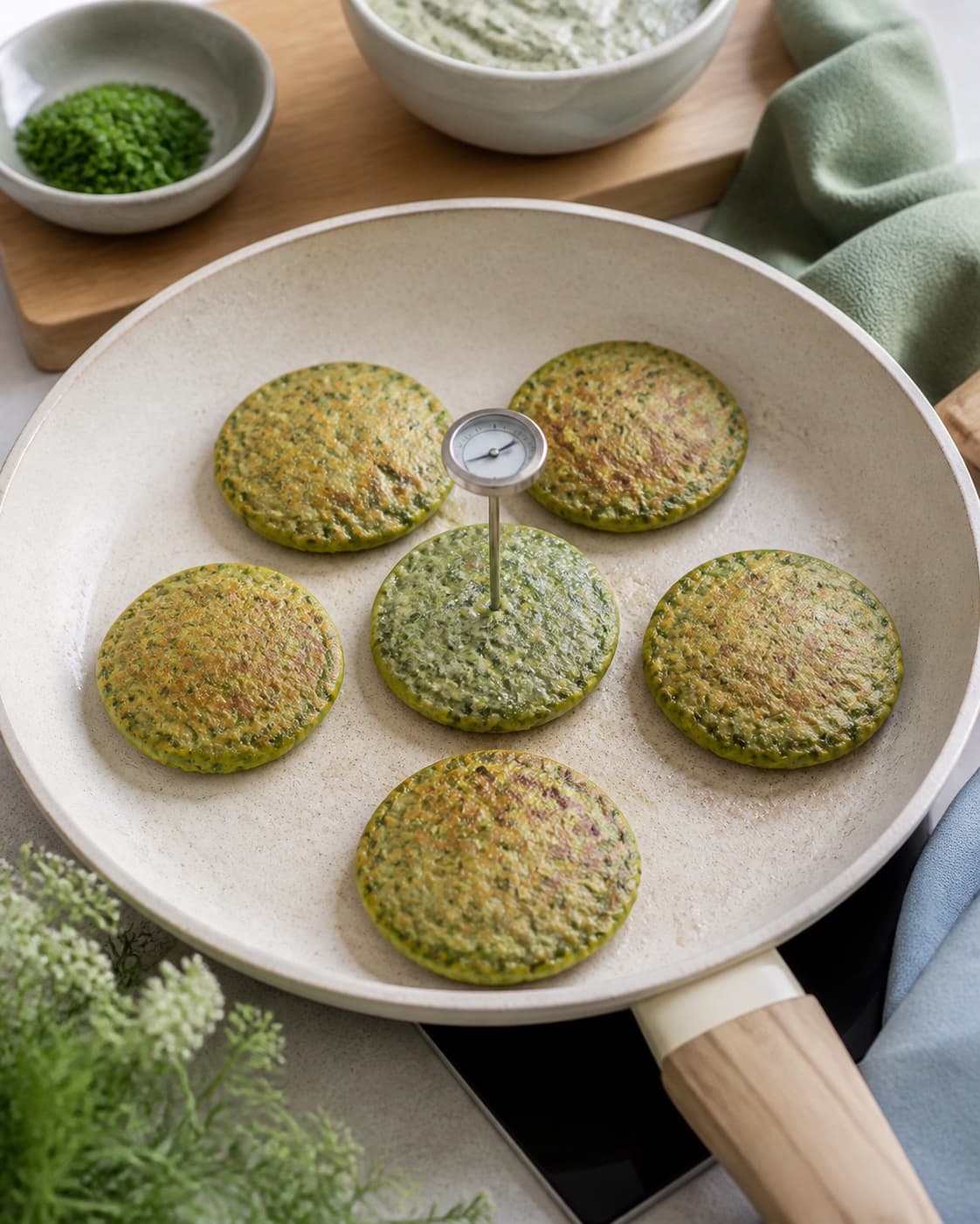 små runde grønne blinis steger i ens størrelse på en lys pande med faste bobler og et rent termometer i midten af én vendt blini; kontroltegnet er blinierne er 6–7 cm, let gyldne udenpå, grønne og faste i midten og måler mindst 75 °c
