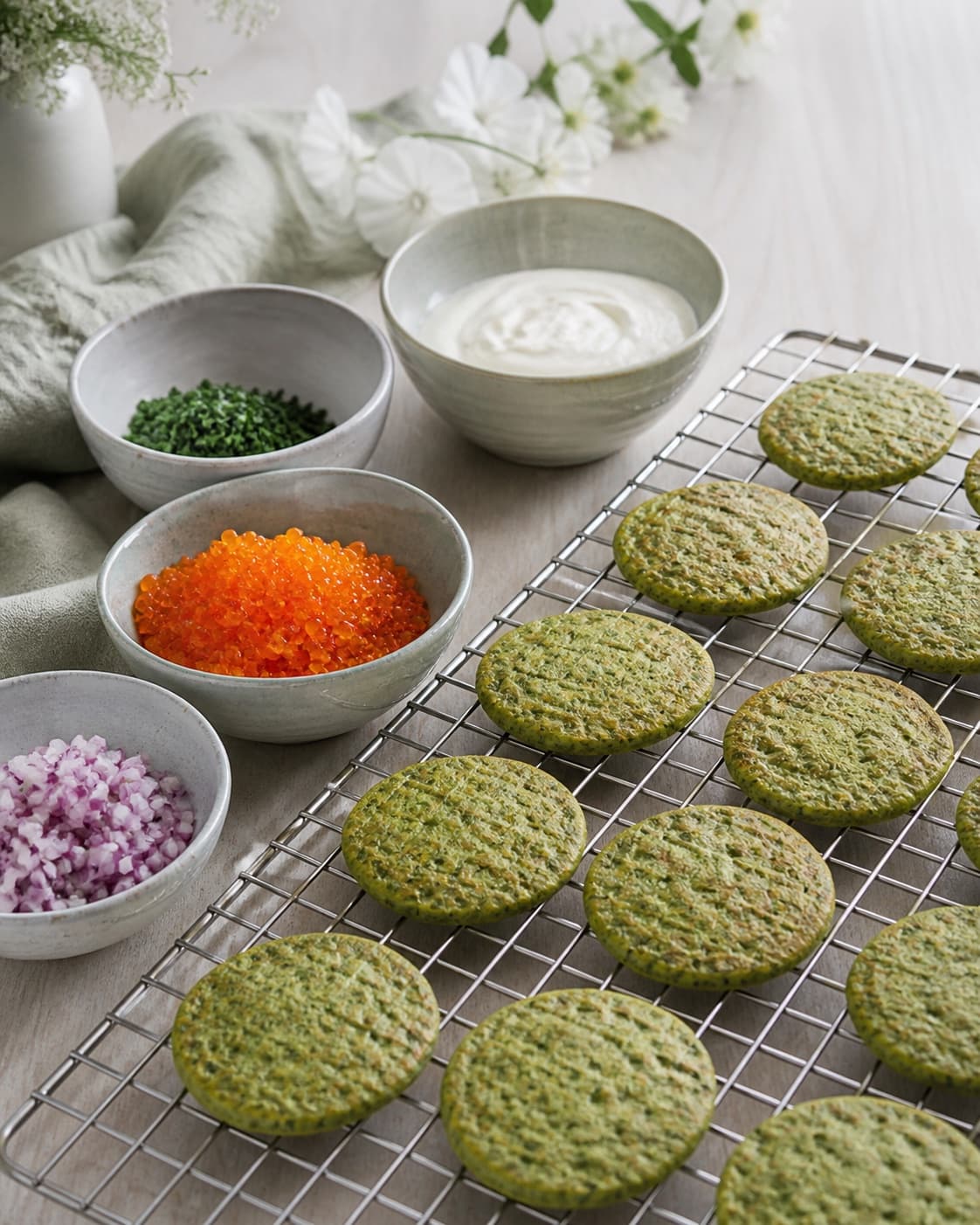 afkølede grønne blinis ligger i ét lag på rist ved siden af separate kolde skåle med creme fraiche orange rogn tørt rødløg og urter; kontroltegnet er blinierne er lune eller kolde, løget er sprødt og tørt, og rogn, creme og urter er højst 5 °c uden væske