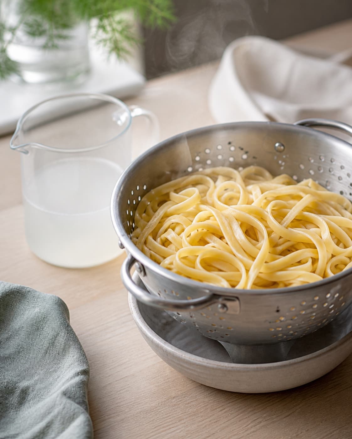 al dente tagliatelle damper i et rent dørslag ved siden af et målebæger let uklart pastavand uden sauce eller laks; kontroltegnet er pastaen er bøjelig med en lille fast kerne, ikke sammenklistret, og pastavandet er let uklart og gemt separat