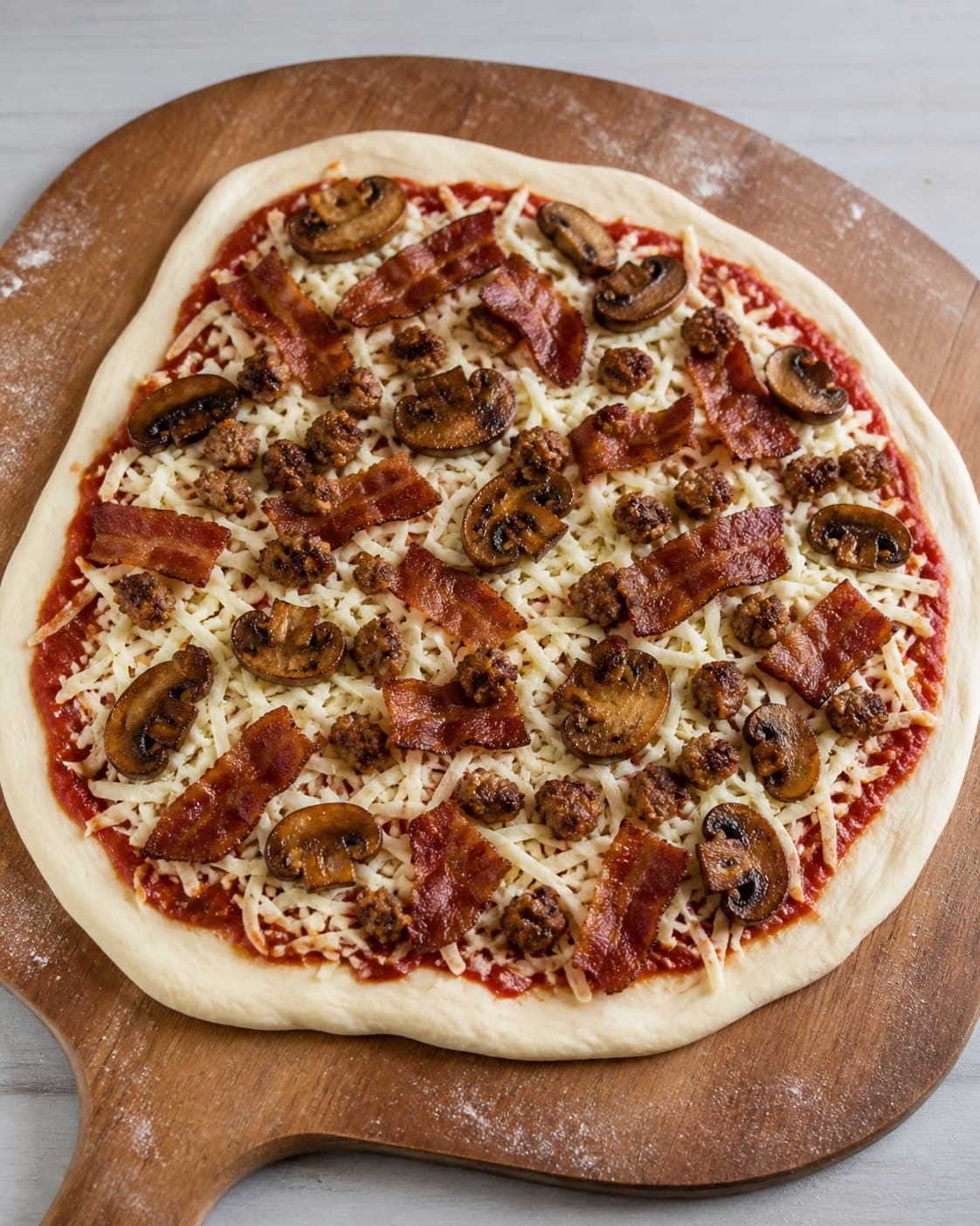 Stretched pizza dough topped with passata mozzarella bacon sausage and mushrooms before first bake under tilberedningen af english breakfast pizza