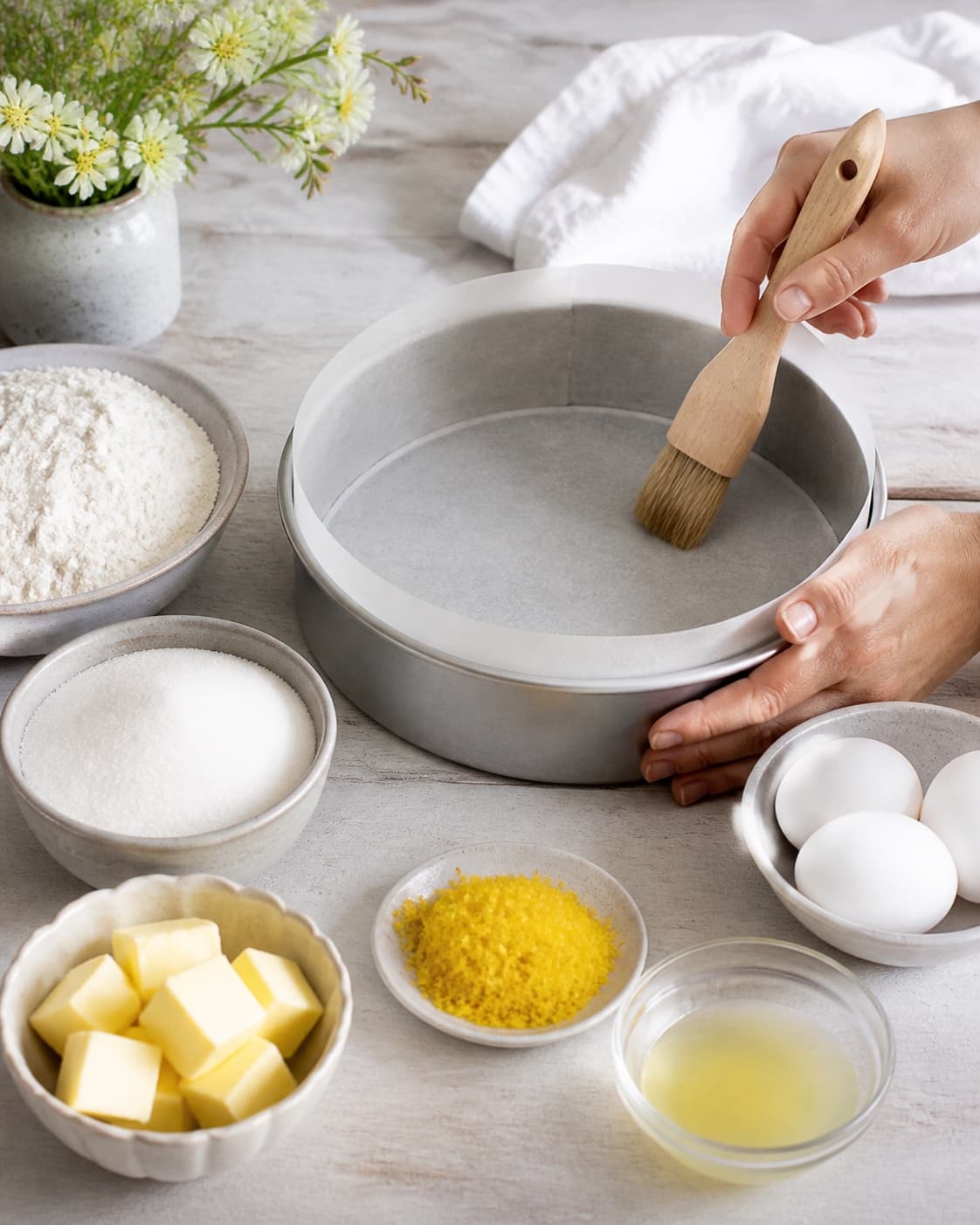 A lined round cake tin beside finely grated yellow lemon zest strained juice flour eggs butter and sugar under tilberedningen af saftig citronmåne med citronglasur