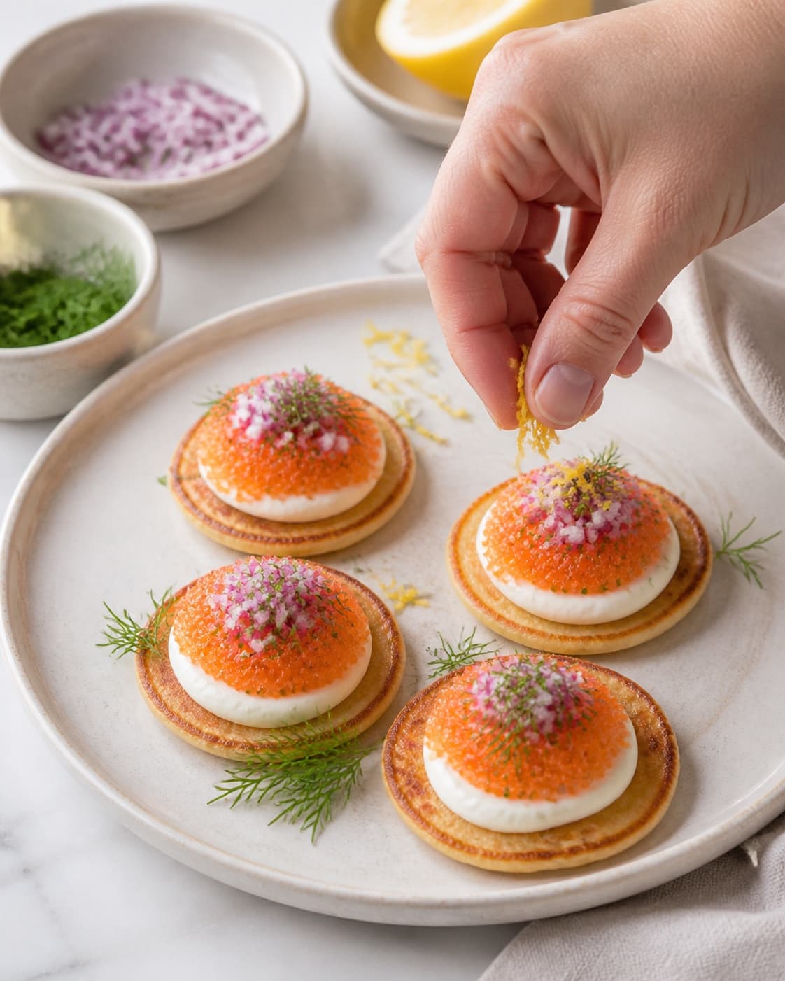 Warm blinis being topped individually with creme fraiche lumpfish roe red onion dill and lemon zest under tilberedningen af blinis med stenbiderrogn