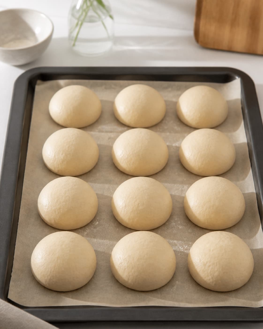 Exactly twelve equal smooth seedless bread rolls being shaped and proofed on a tray under tilberedningen af babyvenlige boller uden kerner