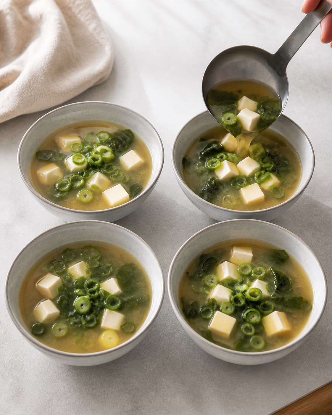 Miso soup with tofu and wakame being ladled into exactly four bowls and topped with spring onion under tilberedningen af misosuppe med tofu og wakame