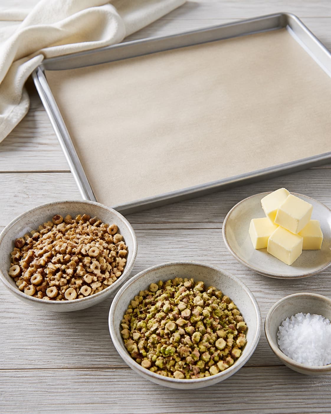 Lightly toasted chopped hazelnuts and pistachios prepared beside an empty parchment-lined tray, butter and flaky salt separate, no caramel under tilberedningen af karamelbrud