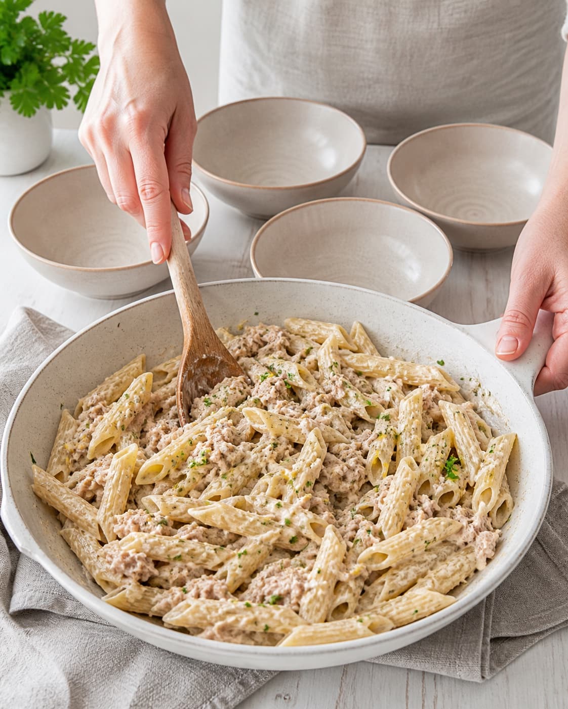 Creamy tuna penne being tossed with parsley and lemon in one pan, glossy sauce clinging to pasta with intact tuna flakes, four bowls ready nearby under tilberedningen af pasta med tunsauce